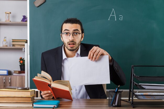 Surprised male teacher wearing glasses holding paper with book sitting at table with school tools in classroom