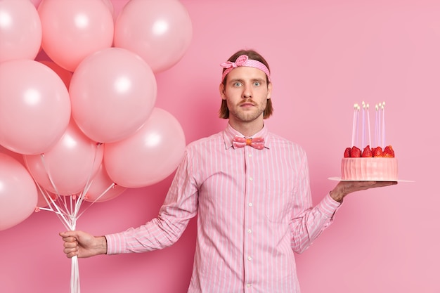 Surprised man celebrates birthday holds bunch of balloons and strawberry cake dressed in formal shirt bowtie shocked to see many guests at party isolated over pink wall