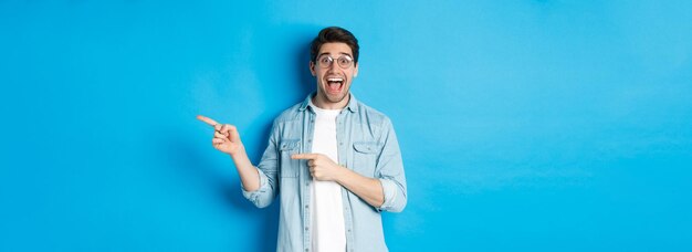 Surprised man in glasses pointing right at copy space showing promo offer on blue background standin