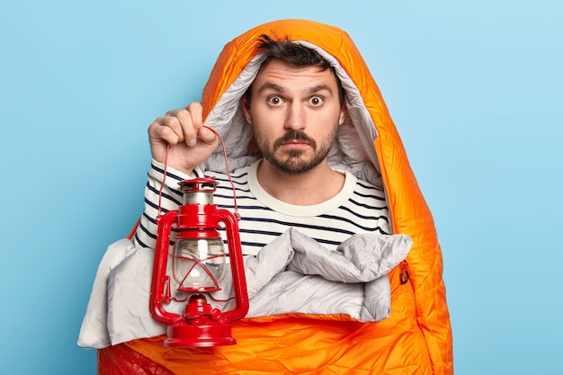 Free photo surprised man has rest, wrapped in sleeping bag, holds kerosene lamp, enjoys recreation in forest, has expedition, poses against blue wall