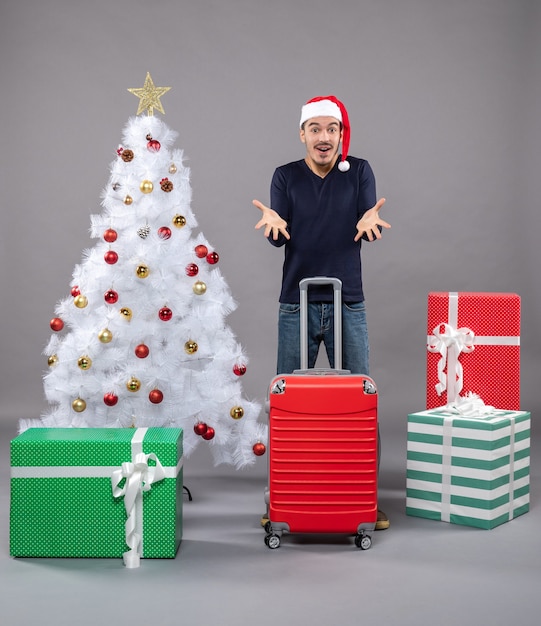 удивленный мужчина держит красный чемодан возле белой рождественской елки с красочными рождественскими игрушками на