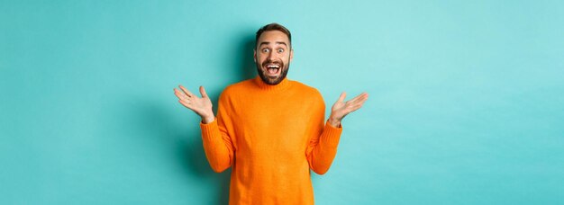 Surprised man raising hands up looking at something amazing standing over turquoise background in wi