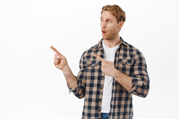 Surprised man say wow looking and pointing left at something interesting checking out event promo staring at advertisement with face showing awe standing over white background