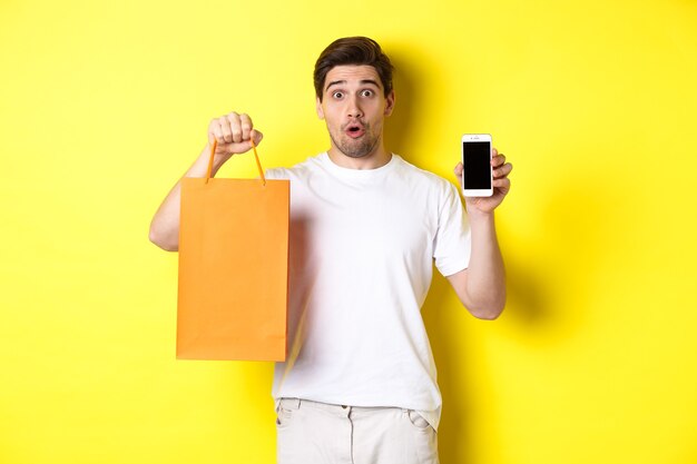 Surprised man showing mobile screen and shopping bag, standing against yellow background