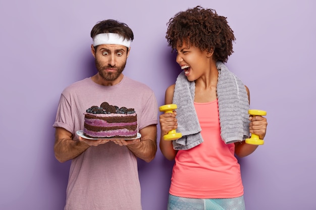 Surprised man stares at baked sweet cake, feels temptation and emotional woman shouts at him, holds yellow dumbbells