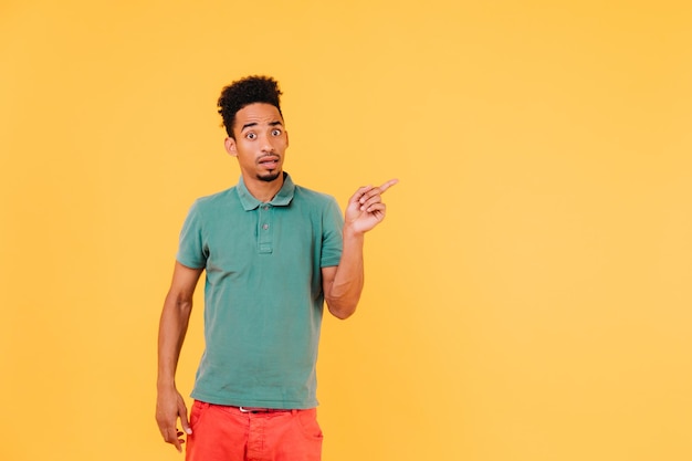 Surprised man in stylish green tshirt pointing finger at something Indoor shot of emotional brunette guy in casual outfit posing on yellow background