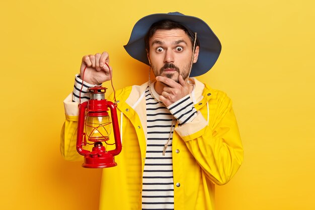 Surprised man traveler keeps hand on chin, wears hat and raincoat, holds little lamp, explores interesting place poses against yellow wall