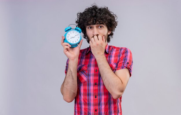 A surprised man with curly hair in checked shirt holding blue alarm clock 