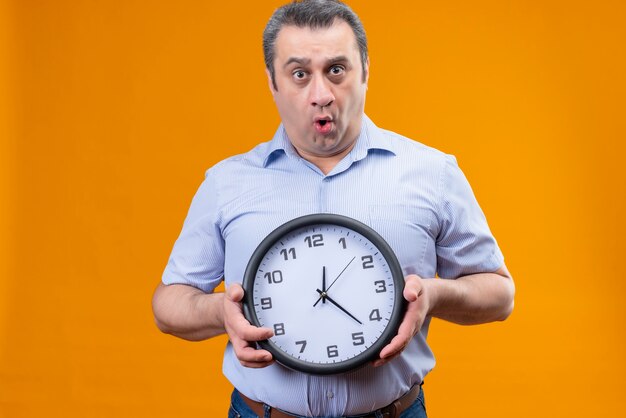 Surprised middle age man in blue striped shirt holding wall clock showing time on an orange background