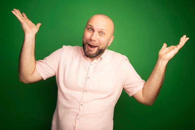 Surprised middle-aged bald man wearing pink t-shirt spreading hands isolated on green