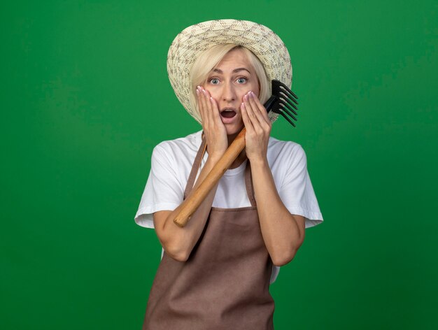 Surprised middle-aged blonde gardener woman in uniform wearing hat holding rake keeping hands on face 