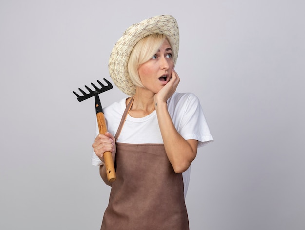 surprised middle-aged blonde gardener woman in uniform wearing hat holding rake putting hand on chin looking at side isolated on white background with copy space