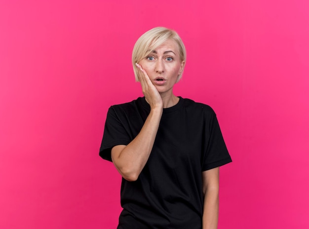 Surprised middle-aged blonde slavic woman keeping hand on cheek looking at camera isolated on crimson background with copy space