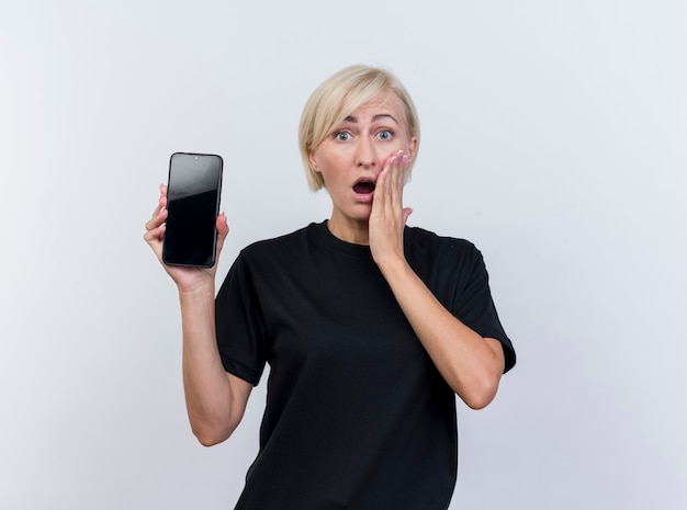 Surprised middle-aged blonde slavic woman showing mobile phone keeping hand on cheek looking at camera isolated on white background with copy space