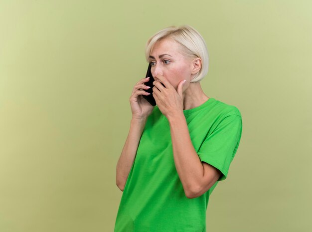복사 공간 올리브 녹색 벽에 고립 된 입에 손을 유지 전화로 이야기 프로필보기에 서 놀란 중년 금발 슬라브 여자