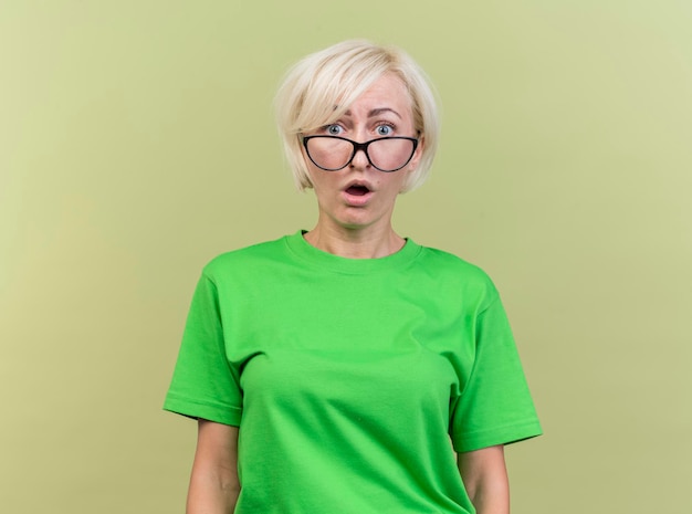 Surprised middle-aged blonde slavic woman wearing glasses looking at front isolated on olive green wall with copy space