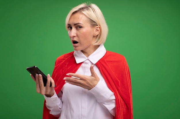 Удивленная блондинка супергероя средних лет в красном плаще держит мобильный телефон, указывая на него рукой