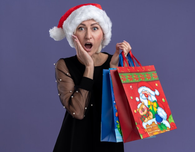 Surprised middle-aged blonde woman wearing christmas hat holding christmas gift bags looking at camera keeping hand on face isolated on purple background