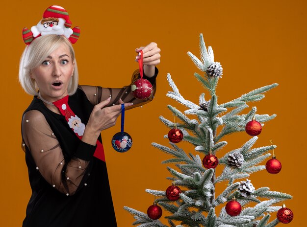 Surprised middle-aged blonde woman wearing santa claus headband and tie standing in profile view near decorated christmas tree holding christmas baubles looking at camera isolated on orange background