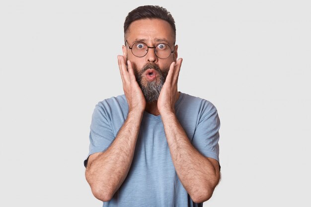 Surprised middle aged Caucasian man with eyewear and rounded lips, dressed gray shirt, stands on white, has astonished expressions, keeps hands on cheeks. People and emotions concept.