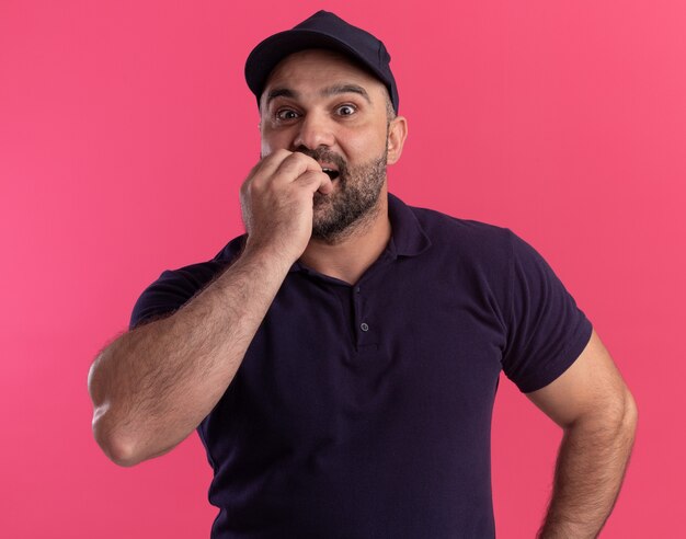 Surprised middle-aged delivery man in uniform and cap bites nails isolated on pink wall