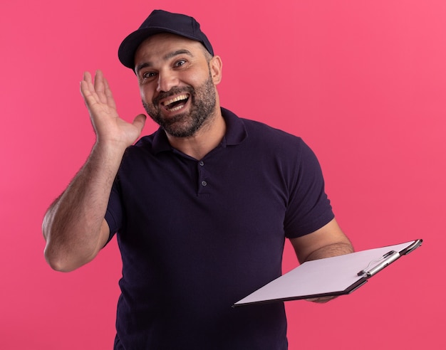 Surprised middle-aged delivery man in uniform and cap holding clipboard isolated on pink wall