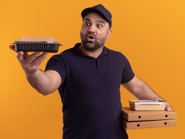 Uomo di consegna di mezza età sorpreso in uniforme e cappuccio che tiene le scatole della pizza e che tiene fuori il contenitore per alimenti isolato sulla parete gialla