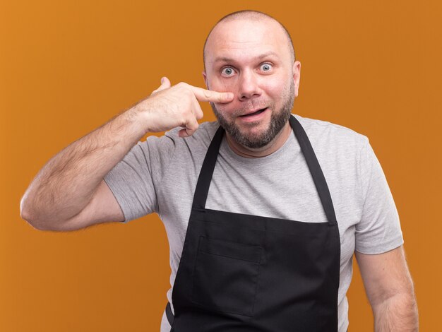 Surprised middle-aged male barber in uniform pulling down eye lid isolated on orange wall