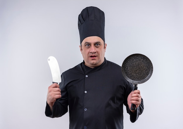 Удивленный мужчина средних лет повар в форме шеф-повара держит сковороду и тесак