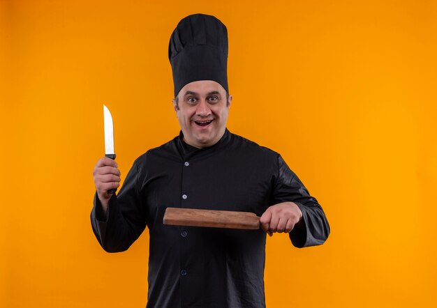 コピースペースと黄色の背景にナイフとまな板を保持しているシェフの制服を着た驚いた中年男性料理人