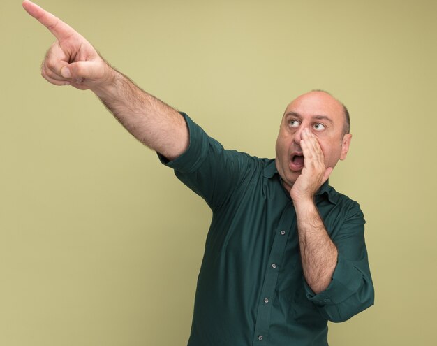 Surprised middle-aged man wearing green t-shirt points at side calling someone isolated on olive green wall