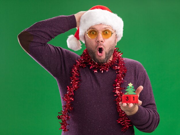 Surprised middle-aged man wearing santa hat and tinsel garland around neck with glasses holding christmas tree toy with date looking at camera