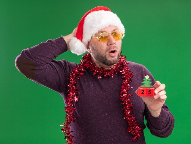 크리스마스 트리 장난감을 들고 안경 목에 산타 모자와 반짝이 갈 랜드를 입고 놀란 중년 남자