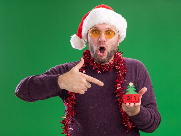 Surprised middle-aged man wearing santa hat and tinsel garland around neck with glasses holding and pointing at christmas tree toy with date looking at camera isolated on green background