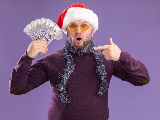 Foto gratuita uomo di mezza età sorpreso che indossa il cappello della santa e la ghirlanda di orpelli intorno al collo con gli occhiali che tengono e che indicano i soldi isolati sulla parete viola