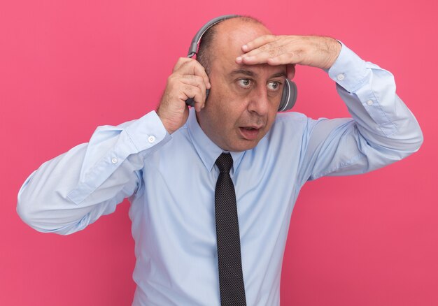 Surprised middle-aged man wearing white t-shirt with tie and headphones looking at distance with hand isolated on pink wall