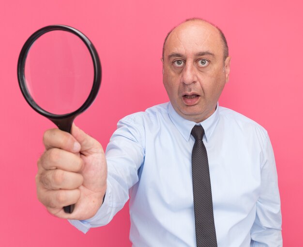 Surprised middle-aged man wearing white t-shirt with tie holding out magnifier  isolated on pink wall