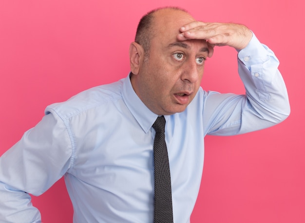 Free photo surprised middle-aged man wearing white t-shirt with tie looking at distance with hand isolated on pink wall