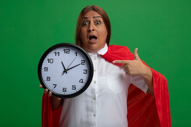 Surprised middle-aged superhero female holding and points at wall clock isolated on green