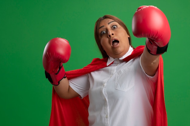 Surprised middle-aged superhero female wearing boxing gloves holding out hands isolated on green