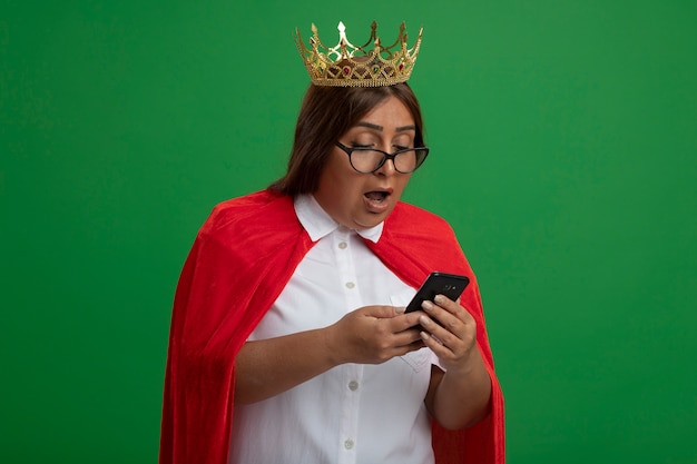 Free photo surprised middle-aged superhero female wearing crown with glasses holding and looking at phone isolated on green