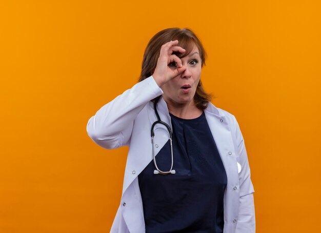 Surprised middle-aged woman doctor wearing medical robe and stethoscope doing ok sign with hand and looking through it on isolated orange wall with copy space