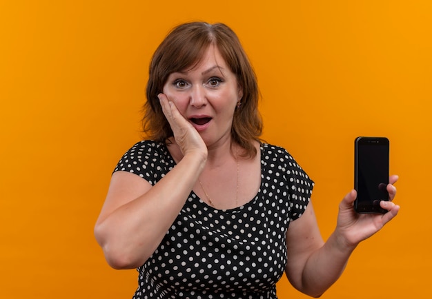 Удивленная женщина средних лет держит мобильный телефон и кладет руку на щеку на оранжевой стене с копией пространства