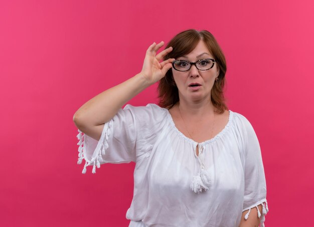 Удивленная женщина средних лет в очках и положив руку на очки на изолированной розовой стене