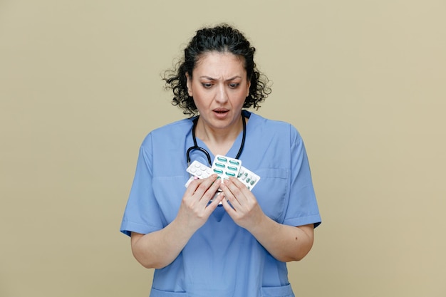 Удивленная женщина-врач средних лет в униформе и со стетоскопом на шее держит упаковки таблеток обеими руками, глядя на них изолированно на оливковом фоне