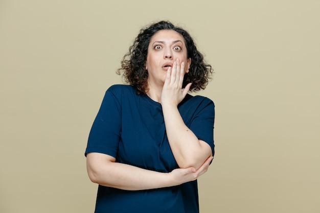 Surprised middleaged woman wearing tshirt keeping hand under elbow and another hand on face looking at camera isolated on olive green background