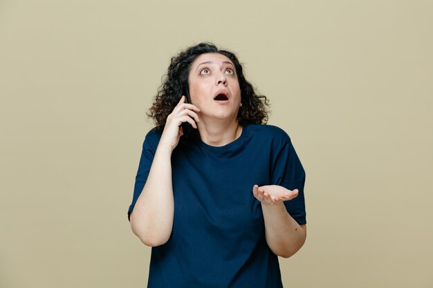 Surprised middleaged woman wearing tshirt looking up showing empty hand while talking on phone isolated on olive green background