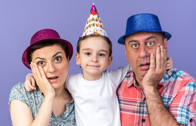 Madre e padre sorpresi con cappelli da festa che mettono le mani sul viso in piedi con il figlio isolato sul muro viola con spazio di copia