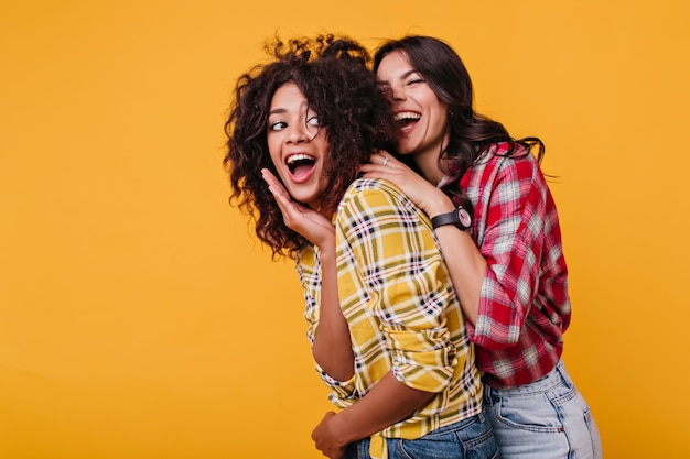 Surprised mulatto covers her mouth from unexpected meeting with her best friend. lady in red shirt with black watch on her hands scared girl from back and laughs