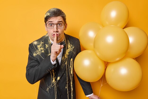 Surprised mysterious adult man makes silence gesture wears spectacles black formal suit smeared with serpentine spray holds bunch of inflated balloons enjoys party celebration Hush be quiet